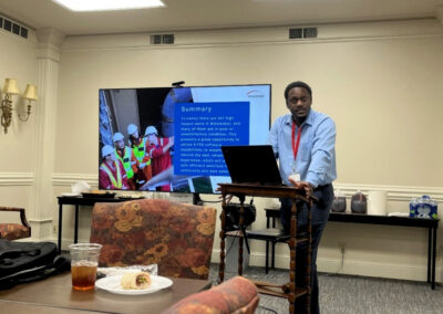 Alex Bohannon presenting a slideshow in a conference room, standing at a wooden lectern with a screen behind him displaying project information.