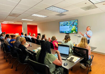 Kevin Day giving a presentation to a group of people seated in a brightly lit conference room. A screen displays the title 'Mapping the Future: Leveraging Data and GIS for Sustainable Community Development.