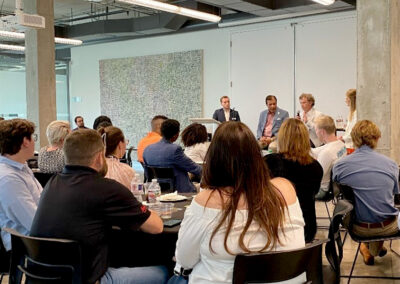 A panel discussion with four speakers seated at the front of a room, addressing an audience of professionals gathered in a modern meeting space.