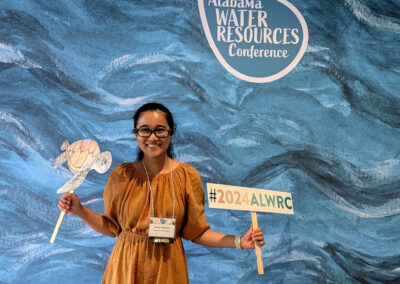 Shrestha standing in front of a blue backdrop with 'Alabama Water Resources Conference' written on it, holding a sign with the hashtag '#2024ALWRC' and a cutout of a turtle.