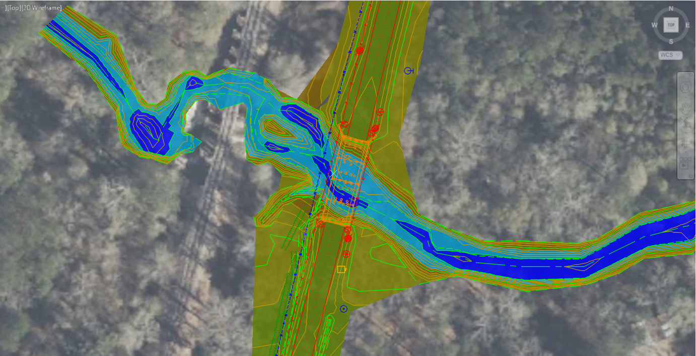 Aerial map overlay showing a detailed hydrological model of a waterway and surrounding area. The model includes multicolored contour lines in blue, green, and yellow, representing elevation changes and water flow patterns. Infrastructure elements such as a roadway and bridge are visible, intersecting the waterway. The background consists of a satellite image of a wooded area.