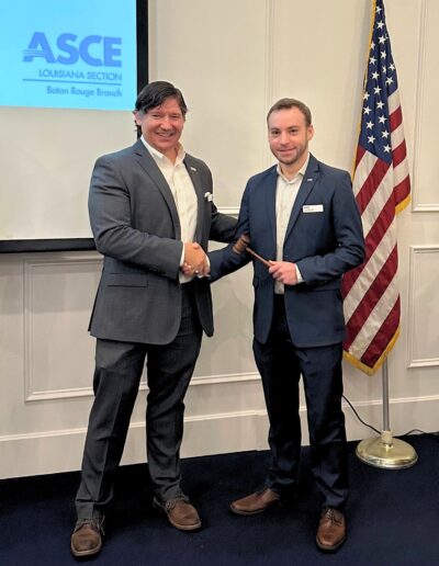 Two men in suits shake hands during a formal event, with one handing over a ceremonial gavel. Behind them, an American flag stands next to a wall-mounted screen displaying the ASCE Louisiana Section Baton Rouge Branch logo.
