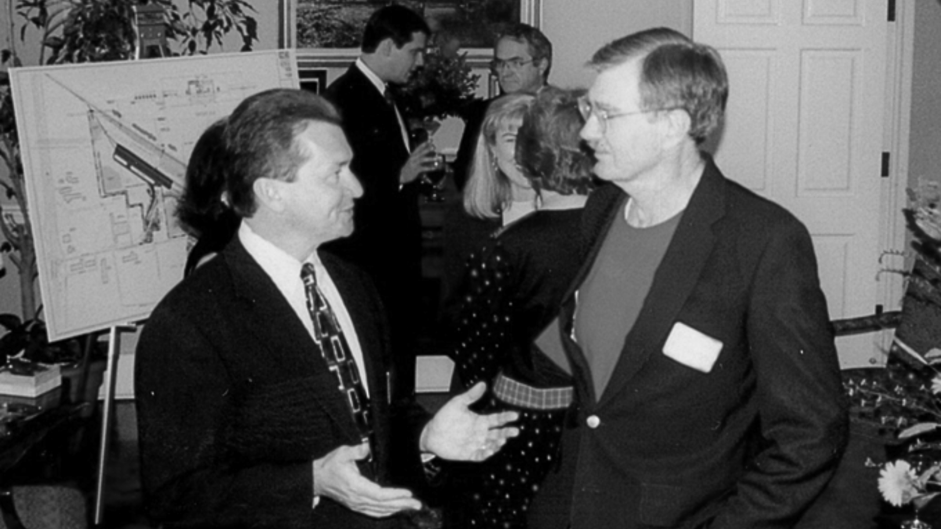 Waggoner - 20th Anniversary in 1996 Black-and-white photo from Waggoner’s 20th anniversary celebration in 1996, showing several people in conversation indoors with a project display board in the background.
